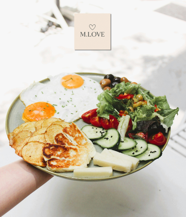 Pessoa segurando um prato com dois ovos estrelados, rodelas de batata frita, queijo grelhado, pedaços de queijo branco, rodelas de pepino com sementes de sésamo, tomates cherry, azeitonas e salada verde.