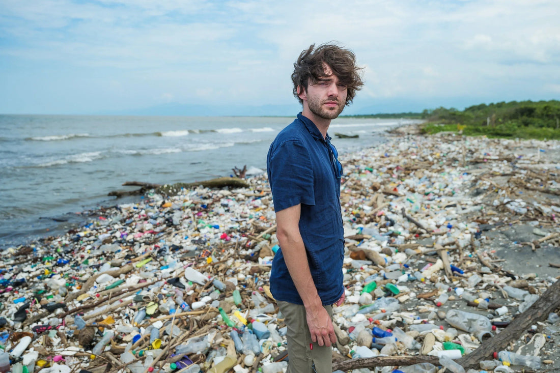 Homem de camisa azul em praia poluída com grande quantidade de lixo plástico e madeira